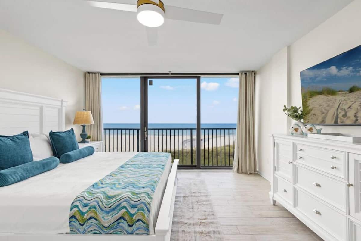 Un dormitorio con una cama grande, ventilador de techo, vista a la playa y cómoda blanca.