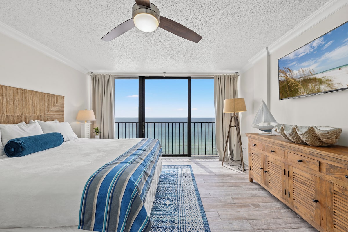 Habitación de hotel con una cama grande, un aparador de madera, un ventilador de techo y vista a la playa.
