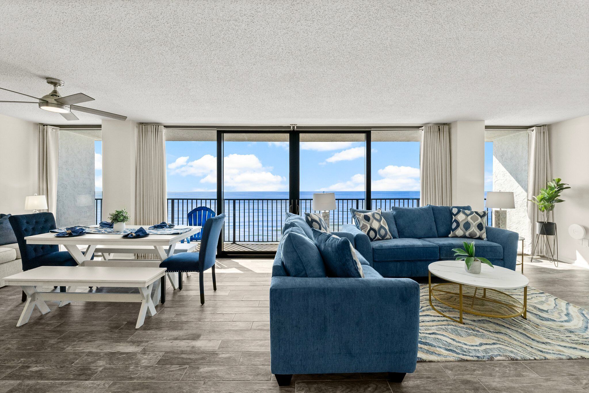 A modern living room with a large window overlooking the sea, a blue sofa, a white coffee table, and a ceiling fan.