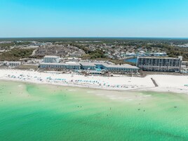Ein Luftbild eines Küstenhotels mit mehreren Gebäuden, einem Strand mit Sonnenschirmen und klarem türkisblauem Wasser.