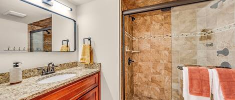 Un baño con lavamanos de encimera de granito, tocador de madera y mampara de ducha con suelo de azulejos.