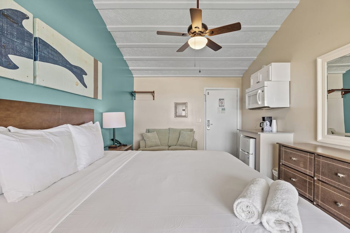 Habitación de hotel con una cama grande, ventilador de techo, microondas, cafetera y baño con lavamanos y espejo.