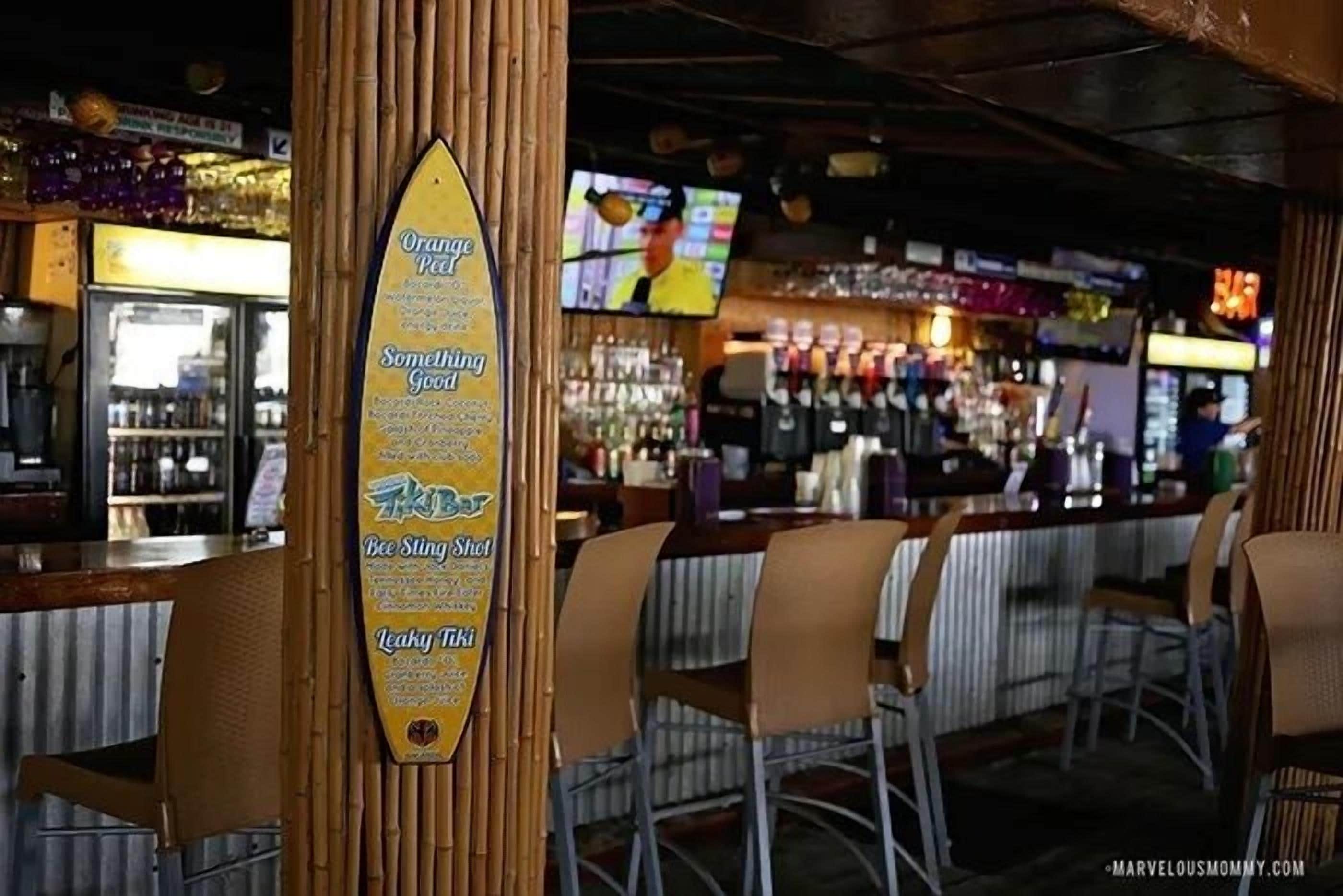 A bar area with a menu board listing drinks like Orange Peel, Somethin' Good, Bee Sting Shot, and Leaky Tiki.