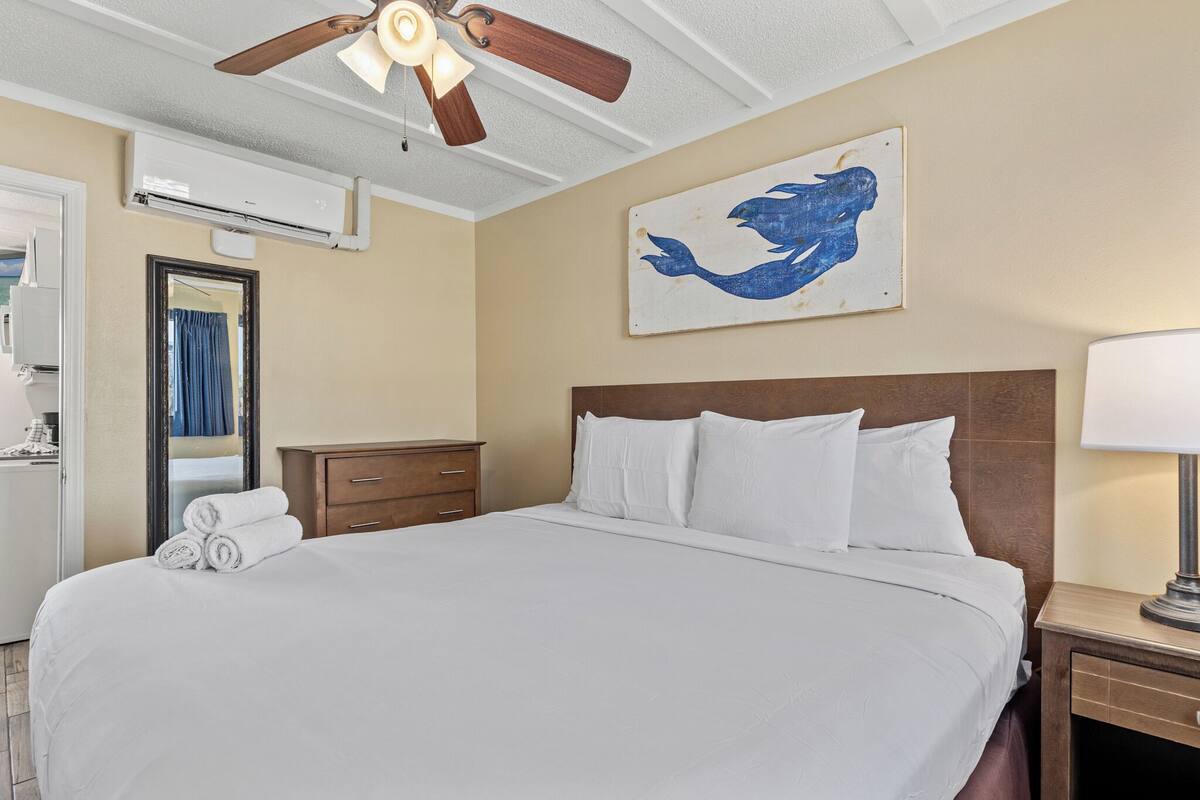 Habitación de hotel con una cama grande, un ventilador de techo, aire acondicionado instalado en la pared, una mesita de noche de madera, un espejo y un cuadro de un pájaro azul.