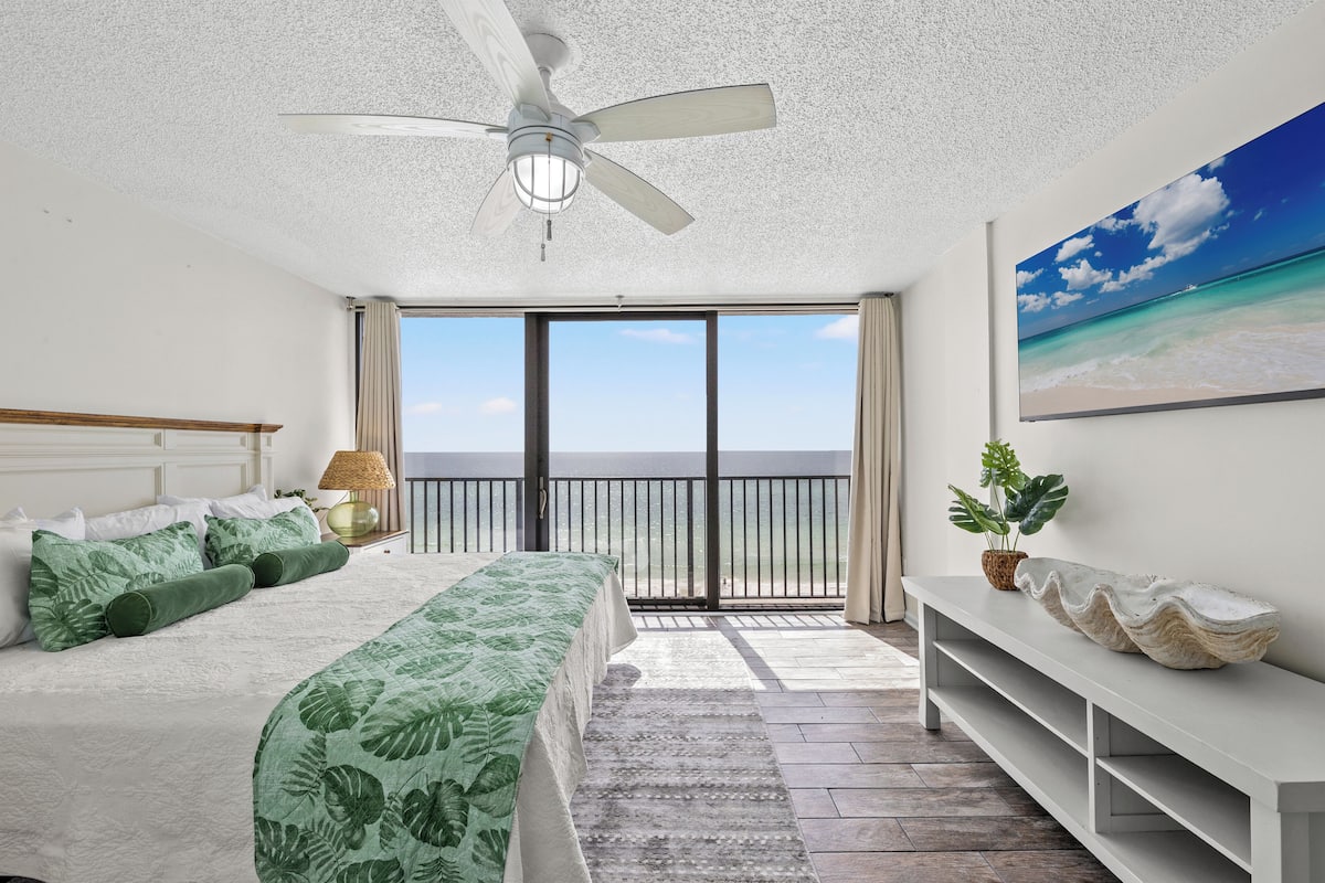 Un dormitorio con cama, ventilador de techo, ventana grande con vista al océano y una consola decorada con objetos.