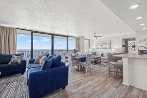 A spacious living area with a large sofa set, dining table, and chairs, a kitchen area, and a balcony with a view.