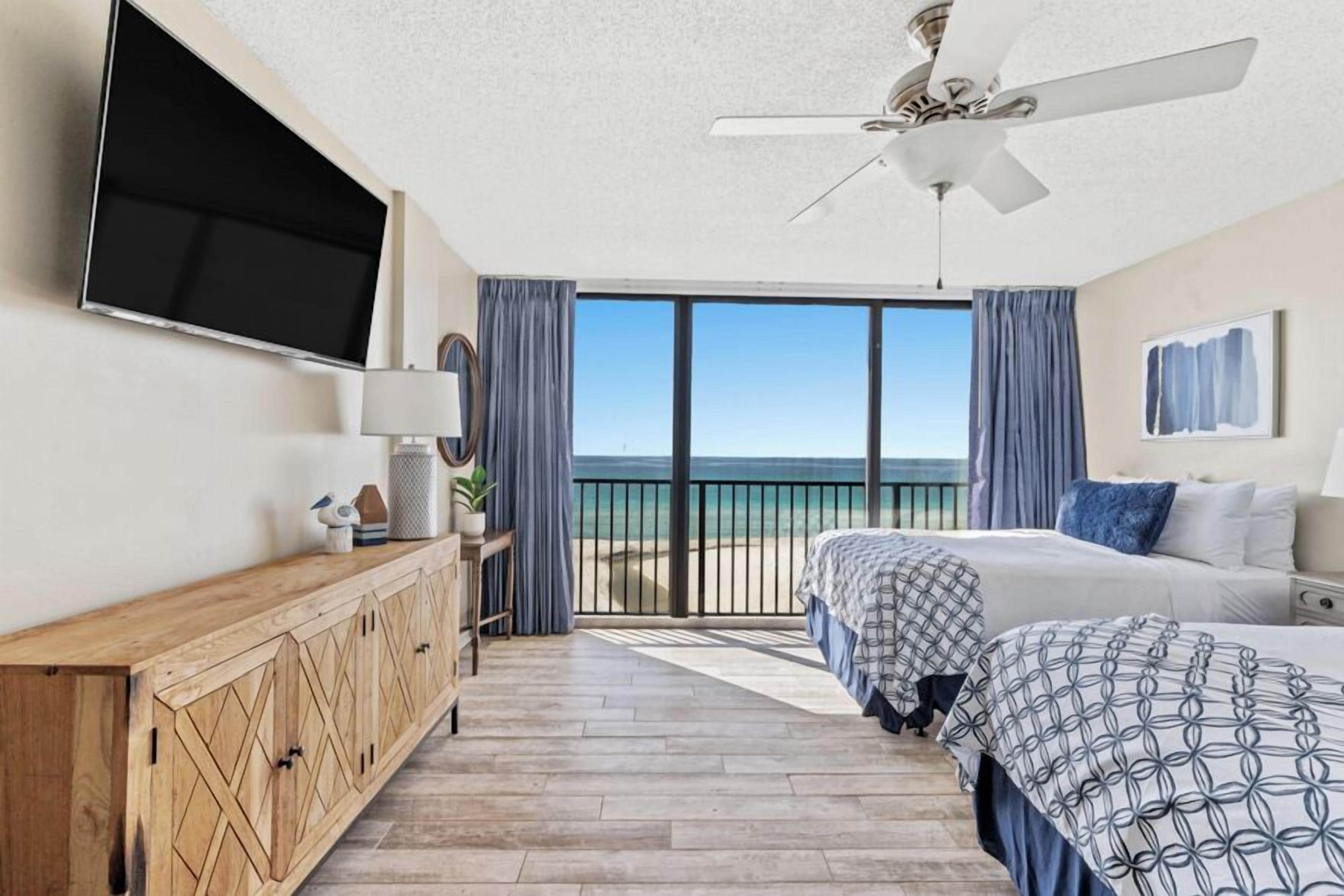 Un dormitorio con una cama grande, ventilador de techo, televisión y vista al océano.