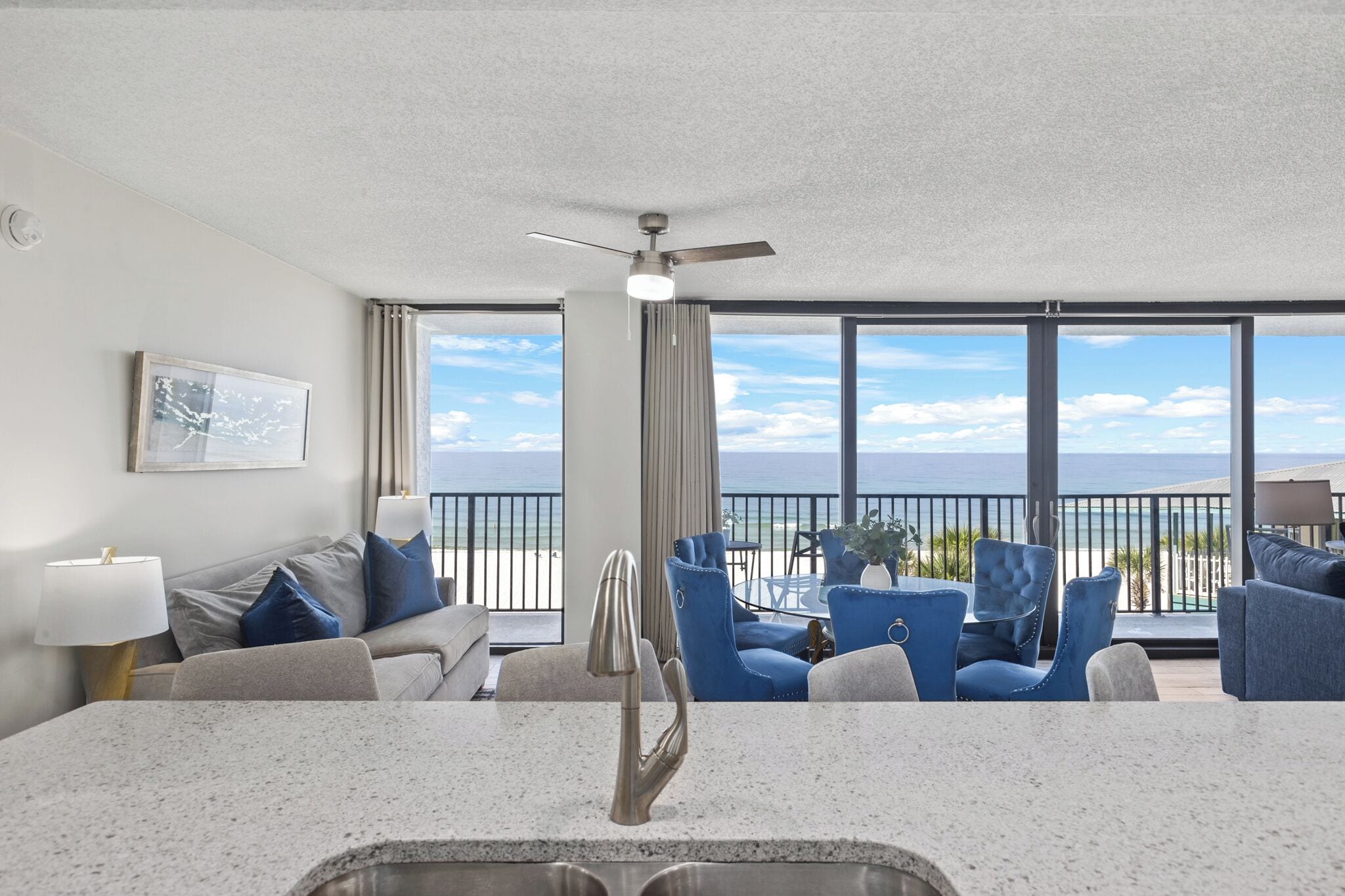 A modern living room with a kitchen area, large windows with a view of the ocean, and a ceiling fan.