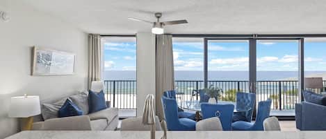 A modern living room with a kitchen area, large windows with a view of the ocean, and a ceiling fan.