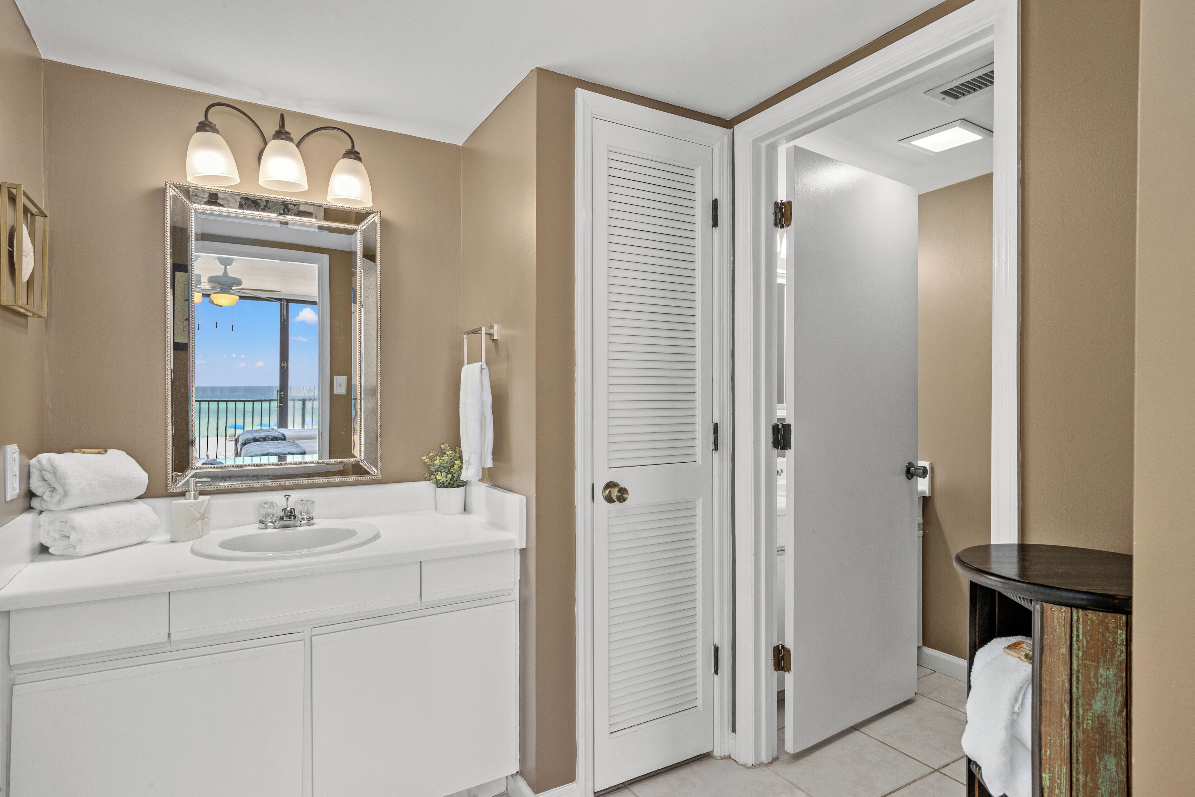 A bathroom with a white vanity, a large mirror, and a view of the ocean through the door.