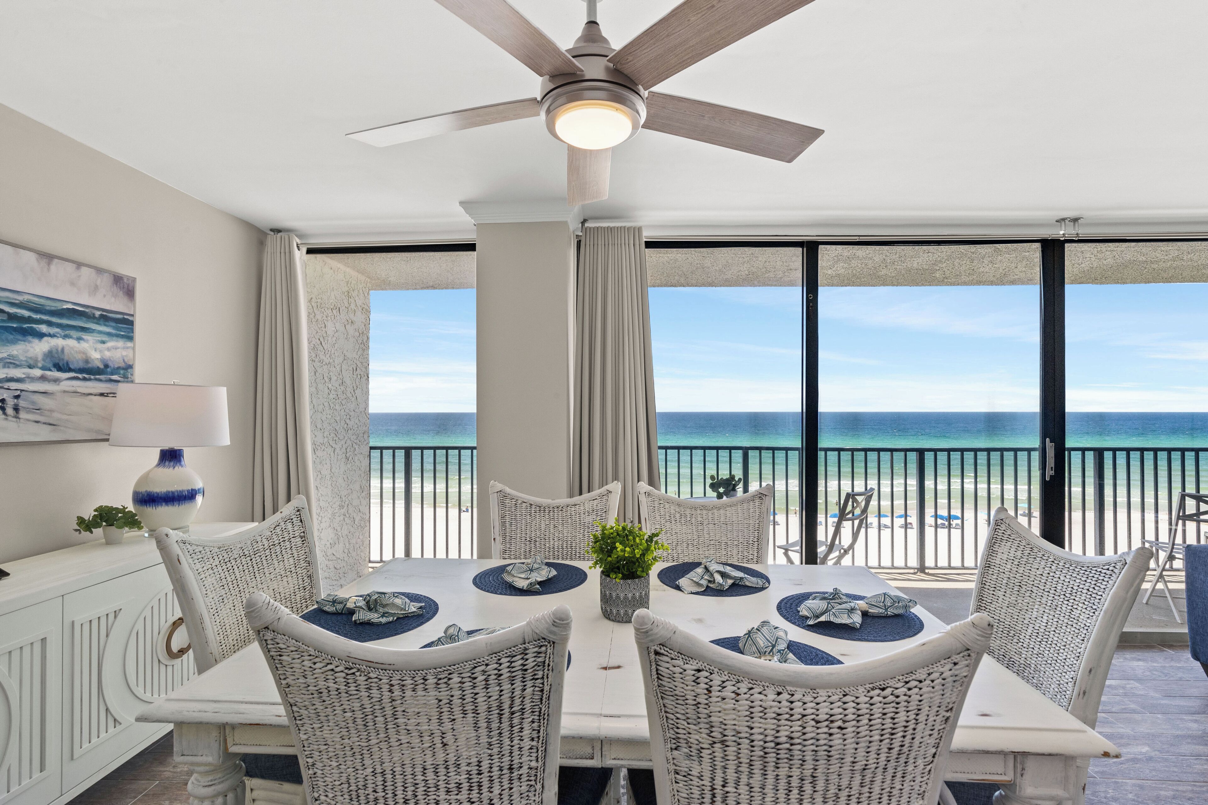 A dining area with a view of the ocean, wicker chairs, a ceiling fan, and a table set for four.