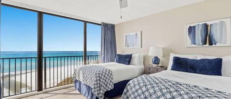 Une chambre d’hôtel avec deux lits, un ventilateur de plafond, une grande fenêtre donnant sur la plage et un balcon avec une balustrade.