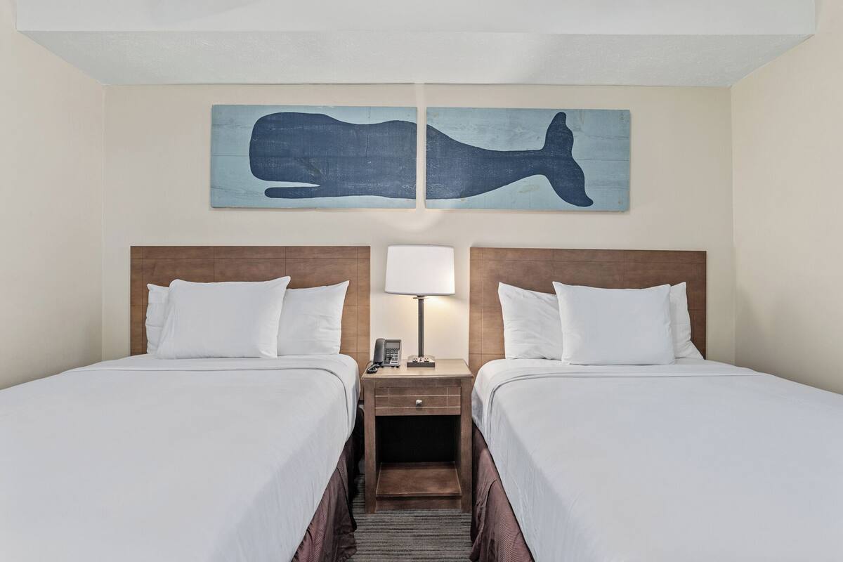 Habitación de hotel con dos camas, una mesita de noche con lámpara y una obra de arte colgada en la pared que representa una ballena.