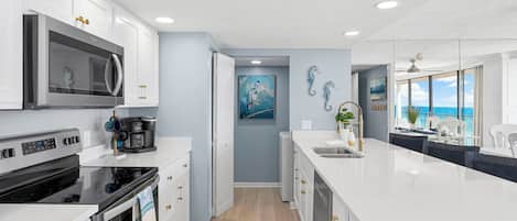 A modern kitchen with white cabinetry, a black stove, and a large island with a sink. There is a painting on the wall and a view of the ocean through large windows.