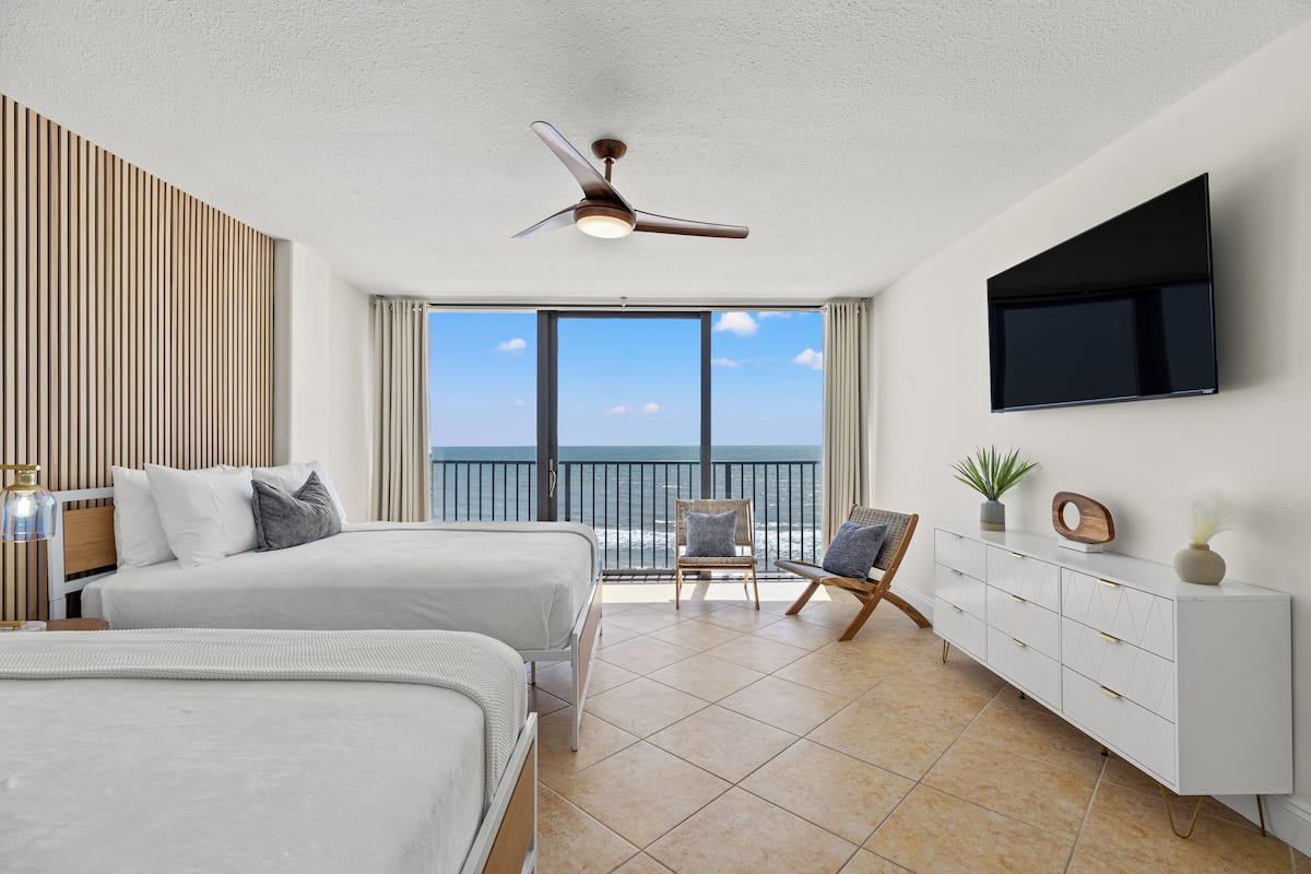 Una habitación de hotel moderna con dos camas, un ventilador de techo, un televisor montado en la pared, un ventanal con vista al mar y un aparador con cajones.
