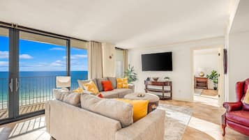 A spacious living room with a large sofa, a coffee table, and a TV. There is a balcony with a view of the ocean.