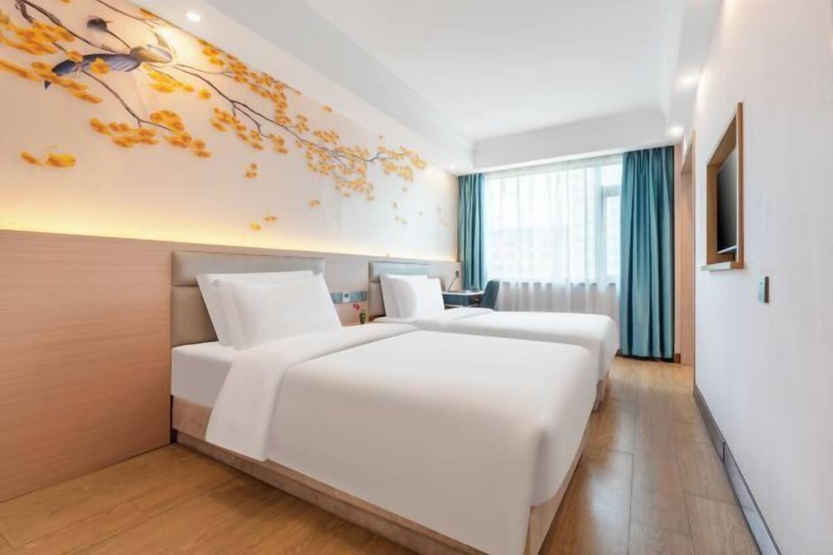 A hotel room with two beds, a wall-mounted artwork, a large window with curtains, and a television on the wall.