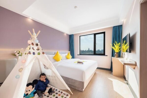 A hotel room with a bed, a window, a desk, and a child playing in a tent.