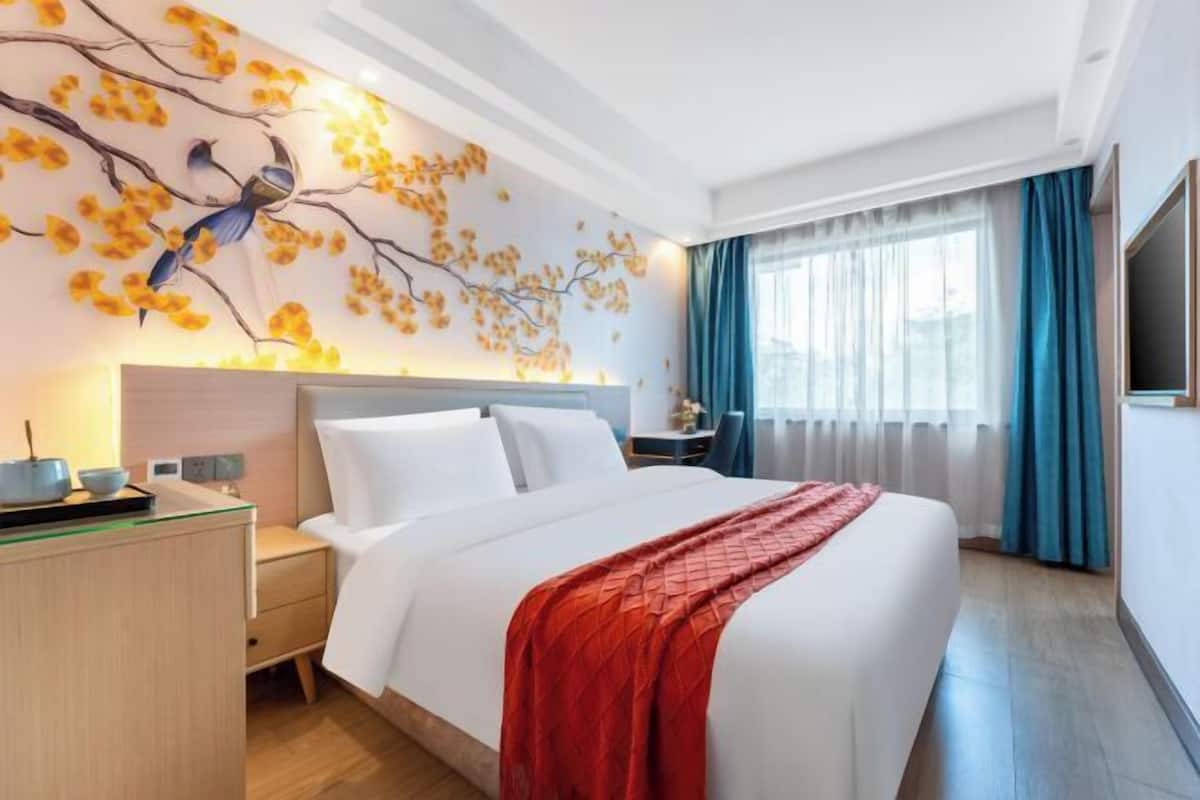 A hotel room with a bed, a nightstand, a desk with a computer, a wall-mounted TV, and a window with curtains.