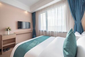 A hotel room with a bed, a TV mounted on the wall, a desk with a vase of flowers, and a window with curtains.