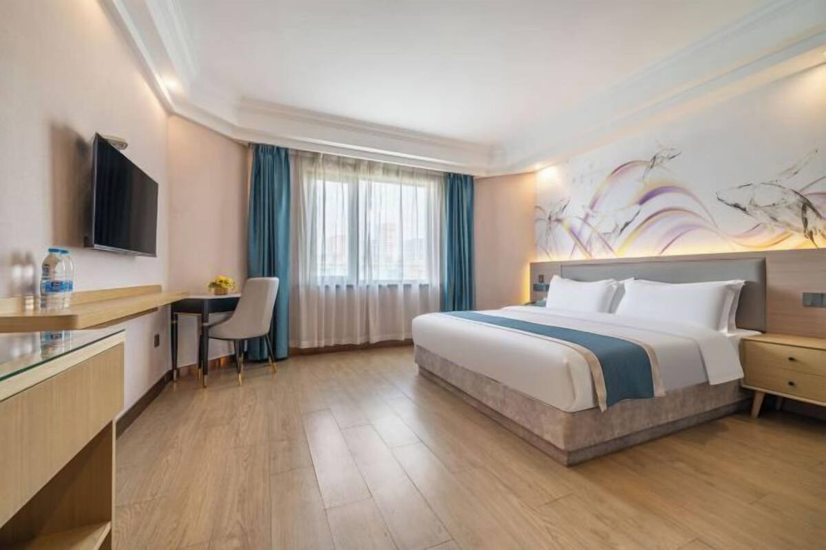 A hotel room with a bed, bedside table, TV, desk, chair, and a painting on the wall.