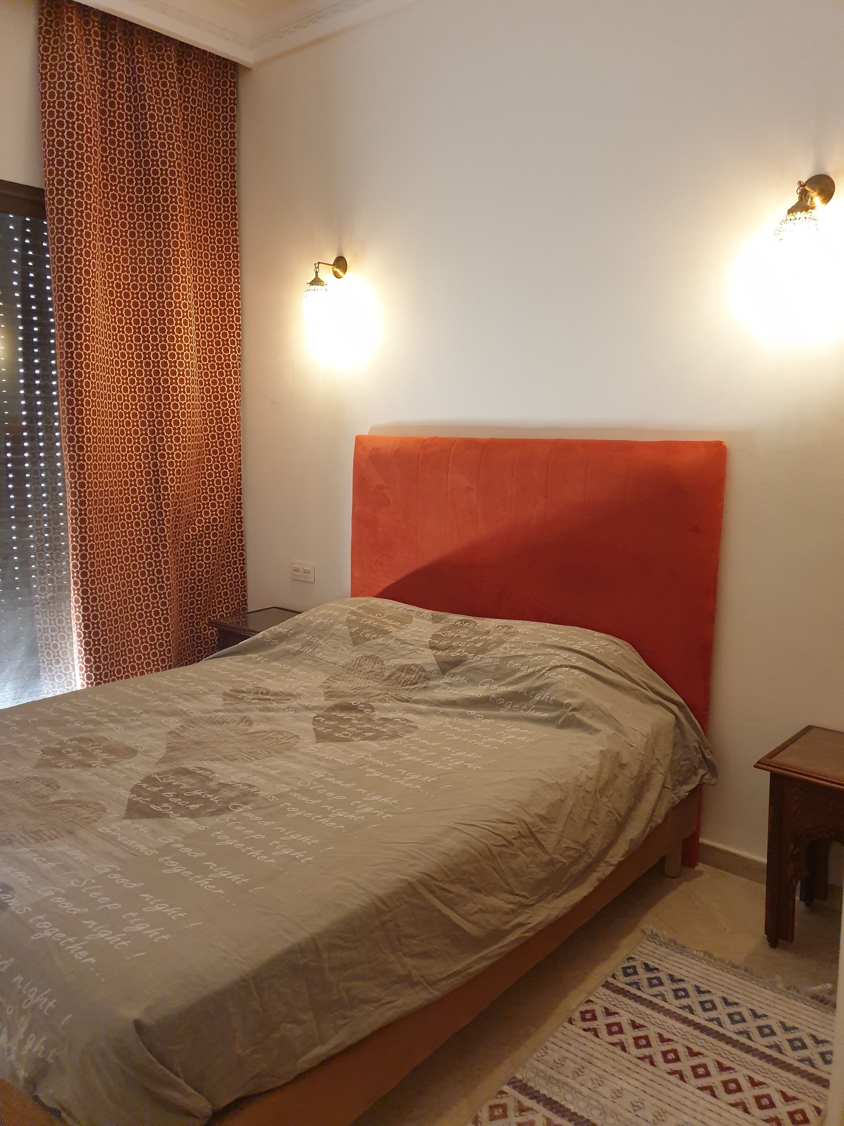Un dormitorio con cama, cortina estampada, dos luces fijadas a la pared y una mesita de noche pequeña.