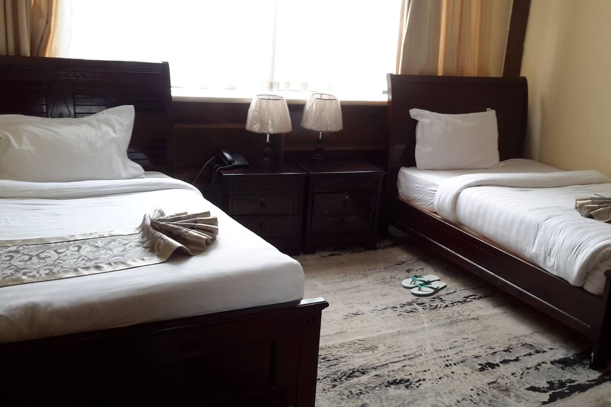 A hotel room with two single beds, a small bedside table with a lamp, and a window with curtains.