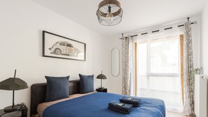 A modern bedroom with a large bed, a nightstand, a lamp, a framed picture on the wall, and a window with curtains.