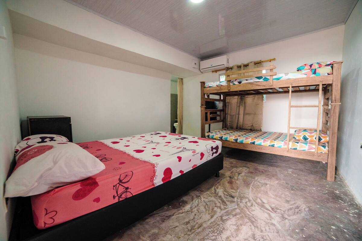 Quarto com duas camas beliche, uma com colcha estampada em vermelho e branco e outra com um design geométrico colorido. Há uma pequena escrivaninha com cadeira e um ar-condicionado fixado na parede.