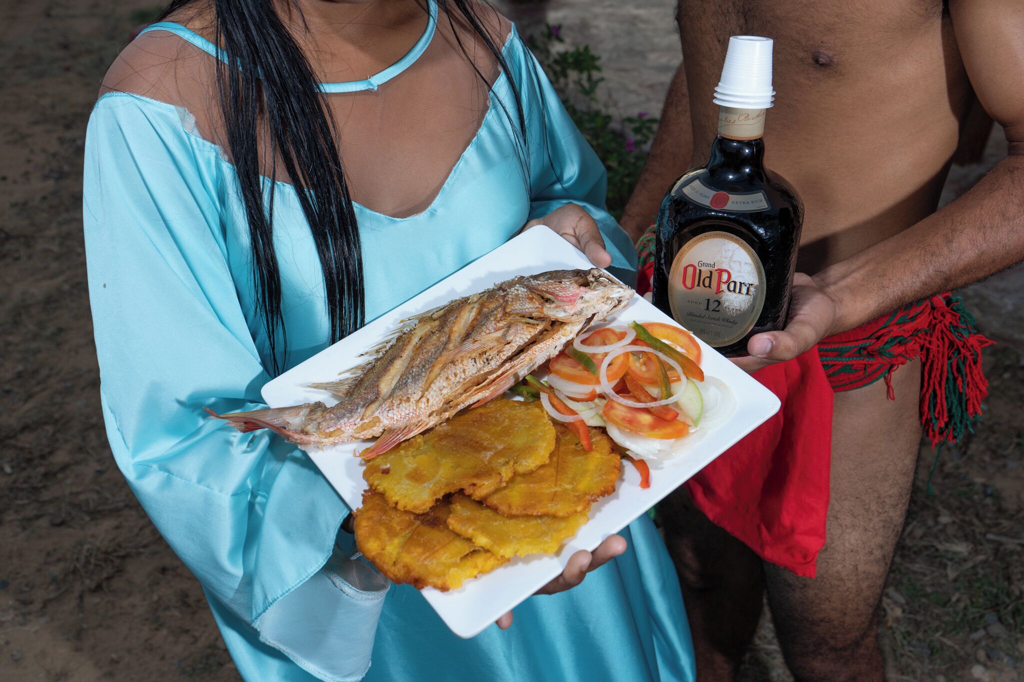 A person holding a plate with fried fish, plantains, and a bottle of Old Parr.
