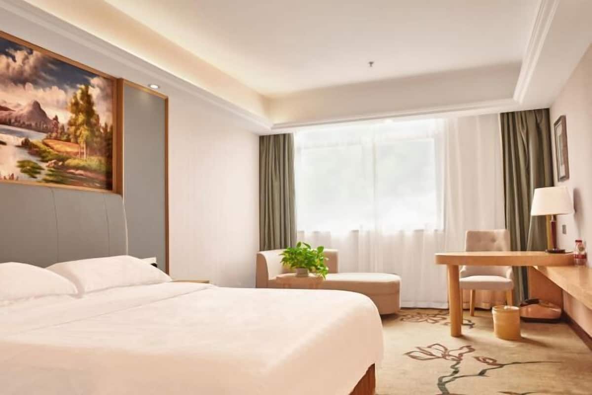 A hotel room with a bed, a desk, a chair, a window with curtains, and a painting on the wall.