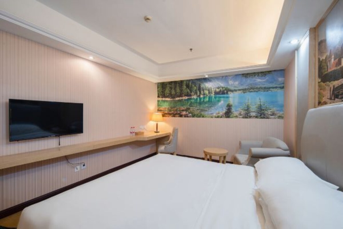 A hotel room with a bed, a TV, a desk with a lamp, a chair, and a painting of a lake.