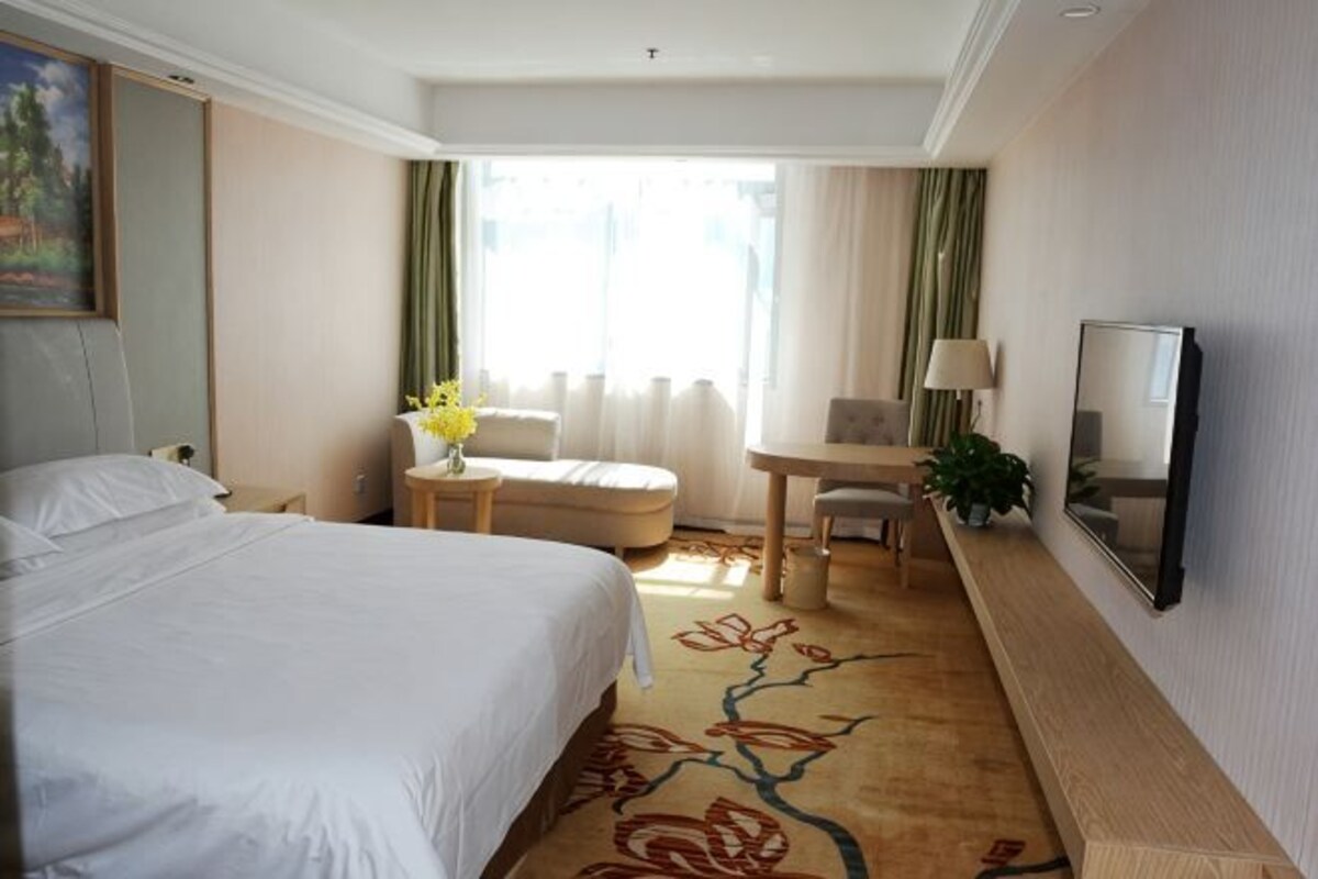 A hotel room with a bed, a desk, a chair, a TV, and a window with curtains.