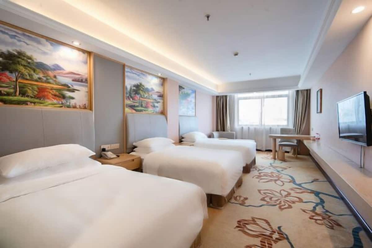 A hotel room with three beds, a flat-screen TV, a desk, and a large window.