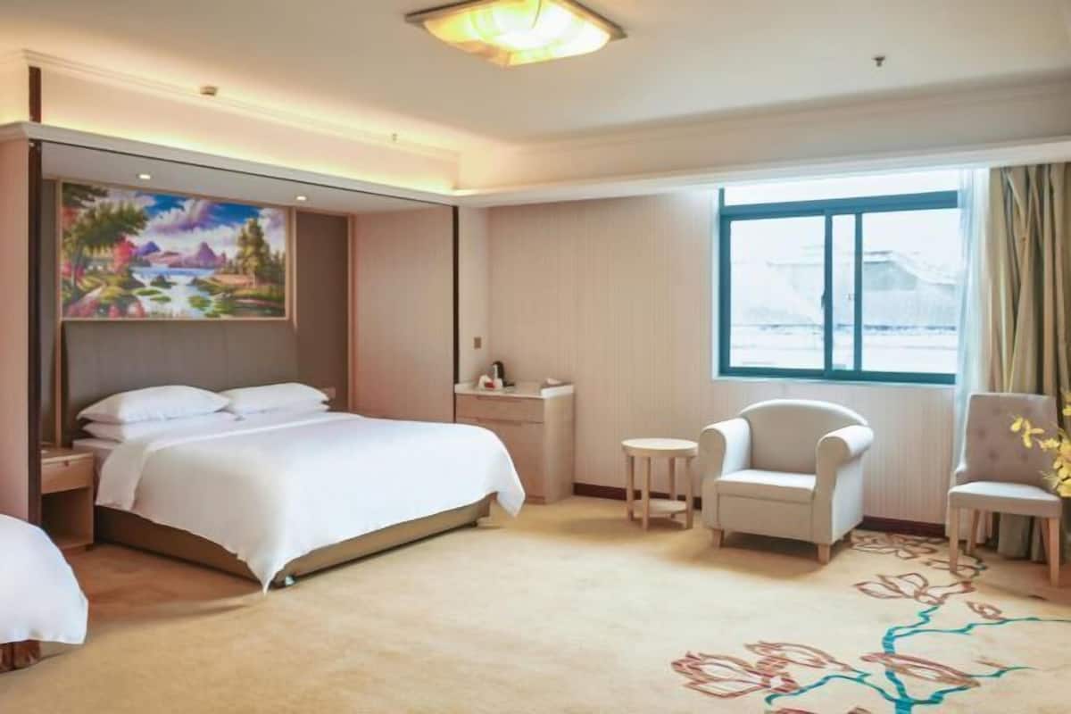 A hotel room with a bed, two chairs, a small table, a painting on the wall, and a window with a view.