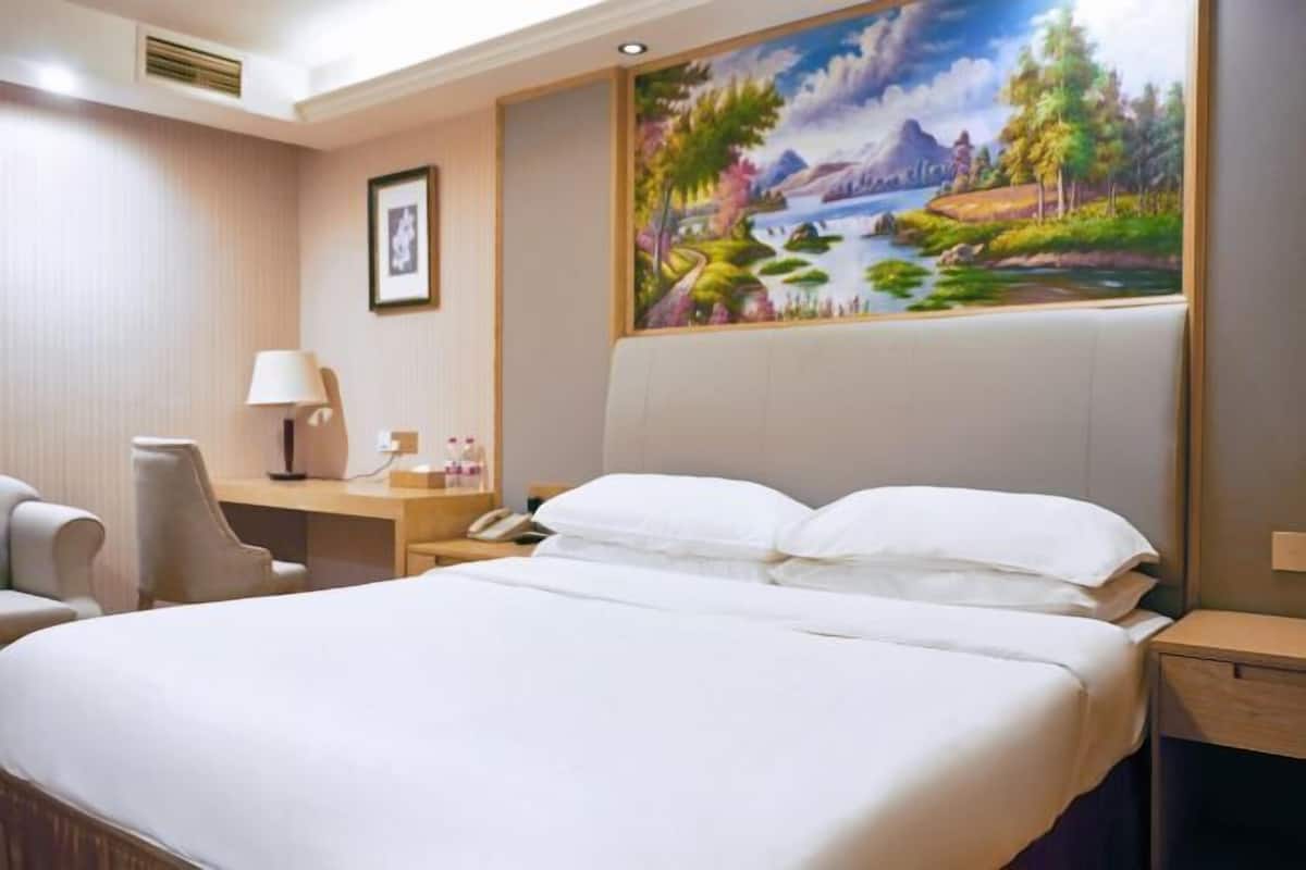 A hotel room with a bed, a desk with a lamp, a chair, and a painting of a landscape.