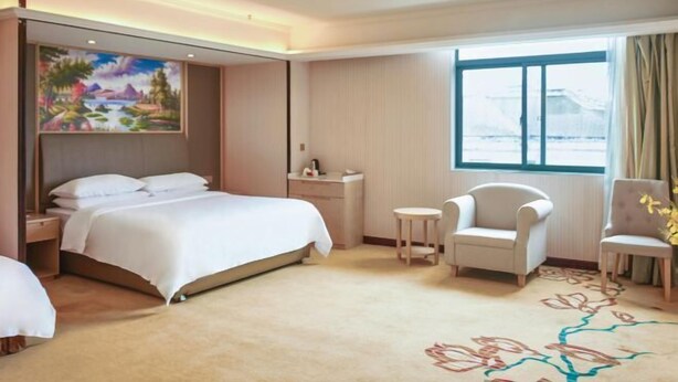 A hotel room with a bed, two chairs, a small table, a painting on the wall, and a window with a view. - Vienna 3 Best Hotel (Shanghai Qiantan Expo Sanlin) (Shanghai)