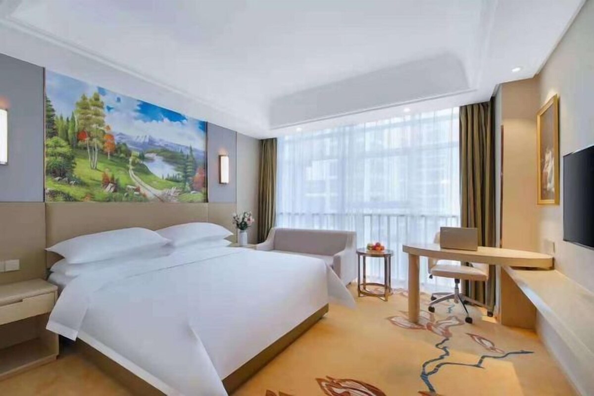 A hotel room with a large bed, a desk, a chair, a small table with a vase, and a painting on the wall.