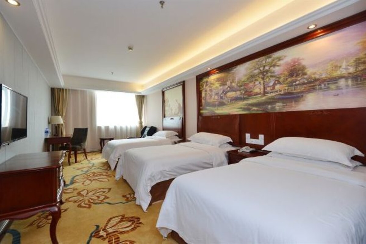 A hotel room with two beds, a desk, a chair, a TV, and a painting on the wall.