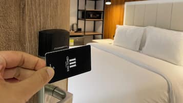 Een hand die een hotel sleutelkort vasthoudt voor een bed met witte lakens en een houten hoofdeinde.