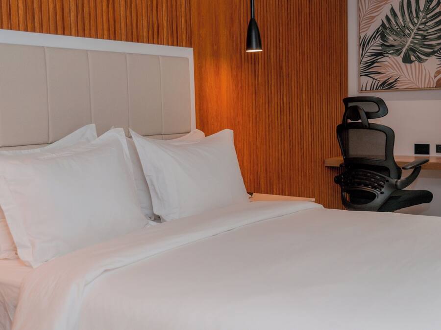 Una cama bien tendida con sábanas blancas, cabecera de madera y una silla negra con auriculares.