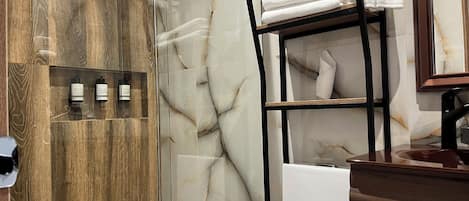 A modern bathroom with a glass shower enclosure, marble walls, a wooden vanity, and a black metal shelf with towels.