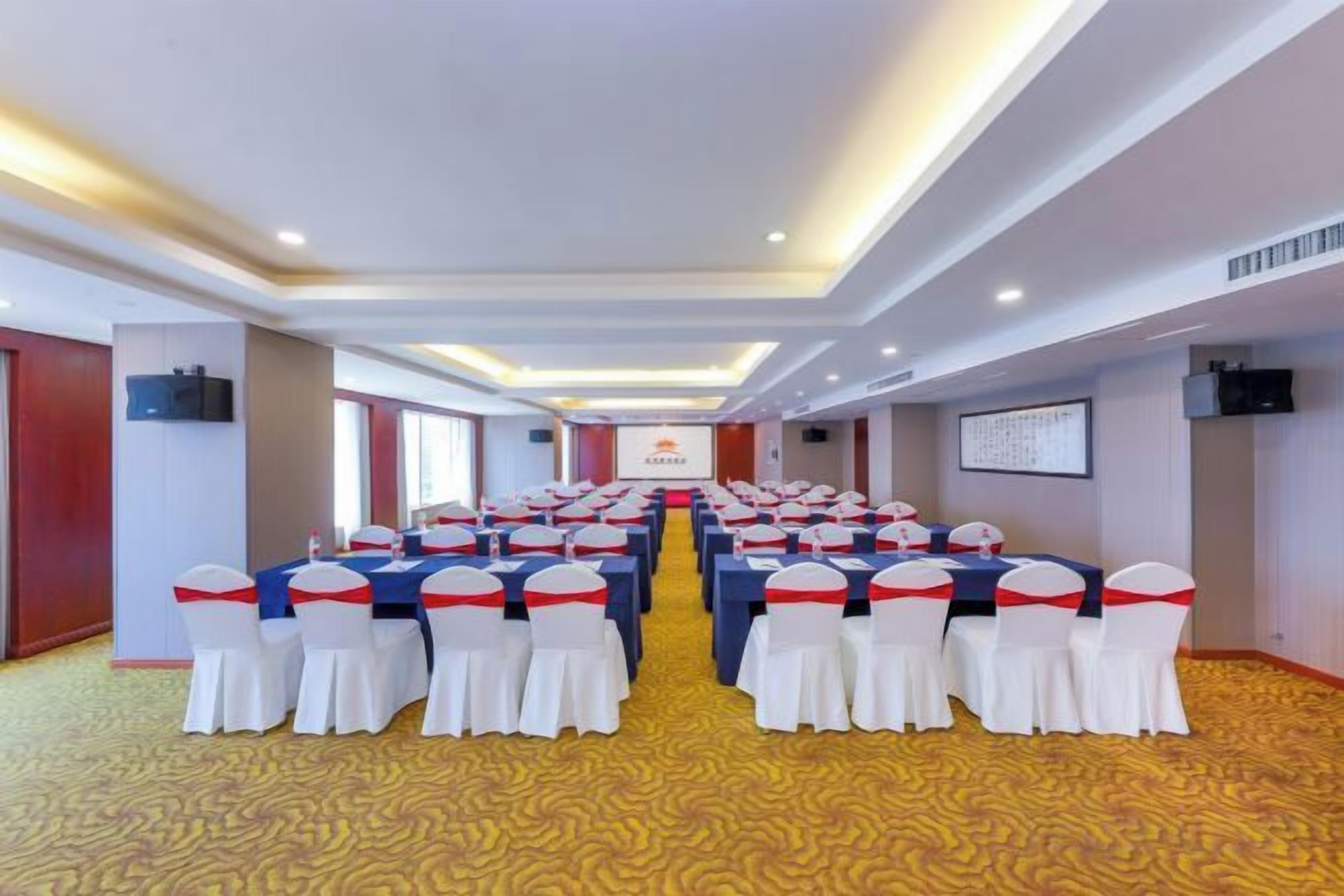 Un salón de conferencias con filas de mesas y sillas, suelo alfombrado e iluminación empotrada en el techo.