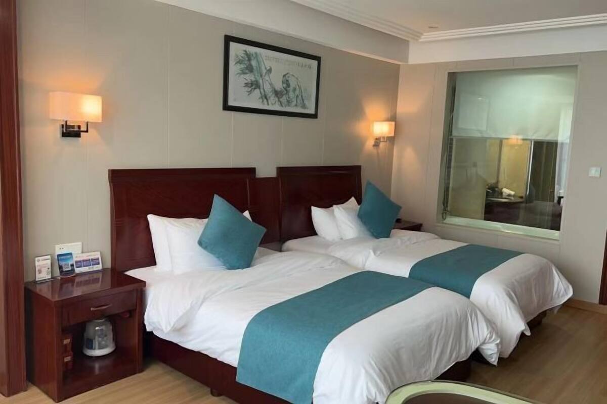 A hotel room with two beds, a nightstand with a lamp, a framed artwork on the wall, and a large window.