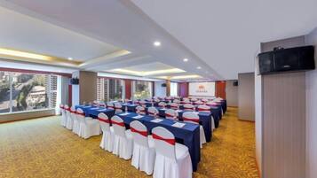 Un salón de conferencias con filas de mesas y sillas, ventanas amplias y vista a edificios.