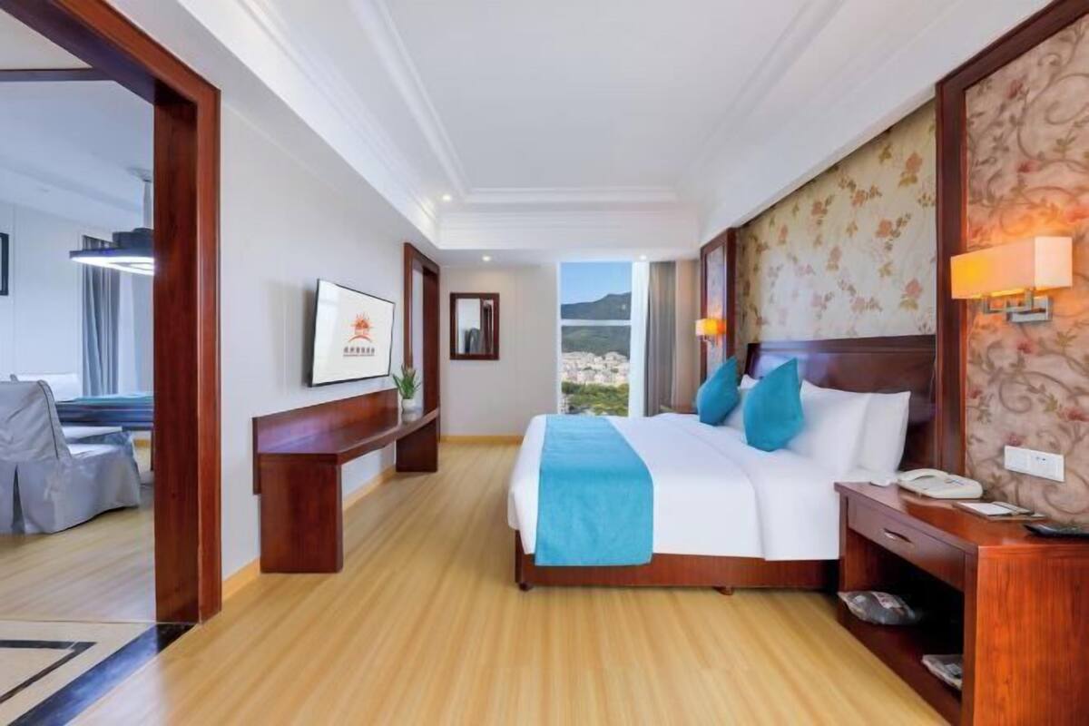 A hotel room with a large bed, wooden headboard, a nightstand, a desk, a chair, and a view of the outdoors.