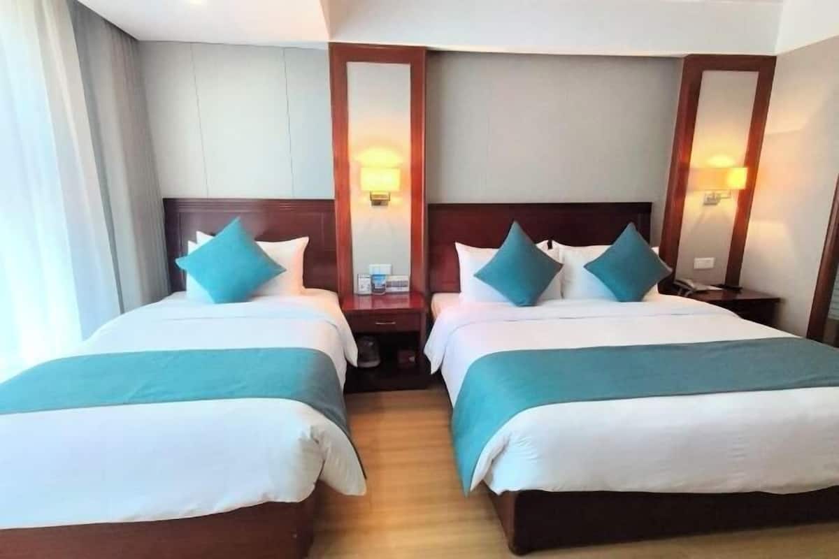 Ein Hotelzimmer mit zwei Betten, jeweils mit einem hölzernen Kopfteil, weißer Bettwäsche und türkisblauen Zierkissen. An den Nachttischen stehen Lampen, und an der Wand ist ein Spiegel angebracht.