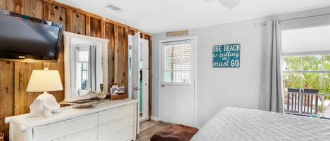 A bedroom with a wooden wall, a TV, a ceiling fan, a bed, a dresser, and a window with a view of greenery.