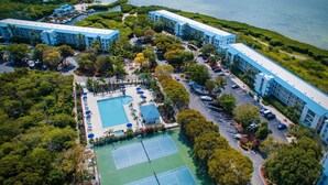 Vue aérienne d’un complexe hôtelier comprenant plusieurs bâtiments, un espace piscine central, des courts de tennis et une végétation luxuriante.