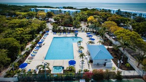 An aerial view of a resort with a large rectangular pool surrounded by lounge chairs and umbrellas, a small building, and a beachfront hotel in the background. - Key Largo 2BR - Partial Ocean Views, Pool & Beach (Tavernier)
